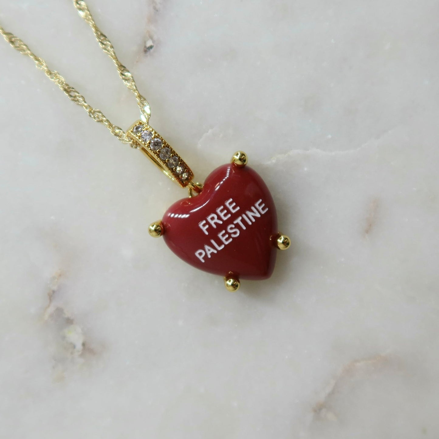 Red heart-shaped pendant with 'FREE PALESTINE' text on a gold chain against a marble background